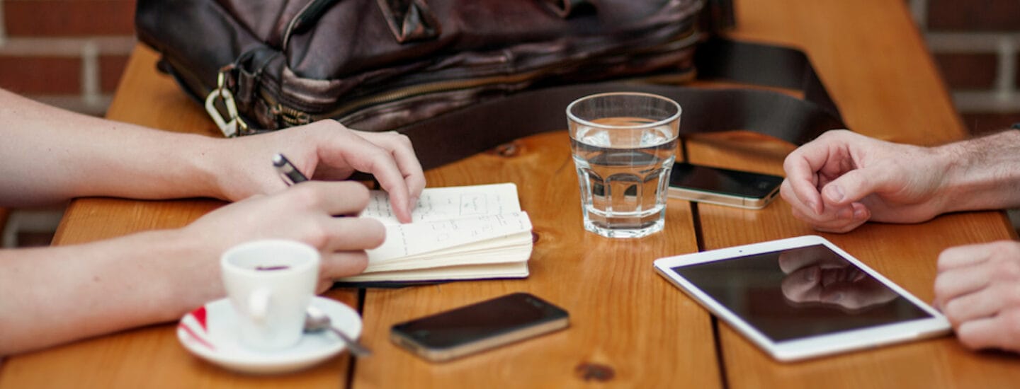 Two people sit at a wooden table, engrossed in conversation. One is writing in a notebook, possibly jotting down notes for rådgivning. Items on the table include a smartphone, tablet, glass of water, cup of coffee, and a leather bag. The setting is casual, likely a café.