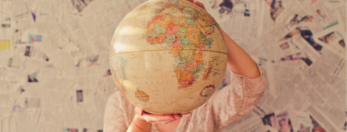 A person holds a large globe in front of their face, obscuring their features like a clandestine figure from the secretariat. The globe displays a vivid map of the world, while the background is a tapestry of overlapping newspaper pages.