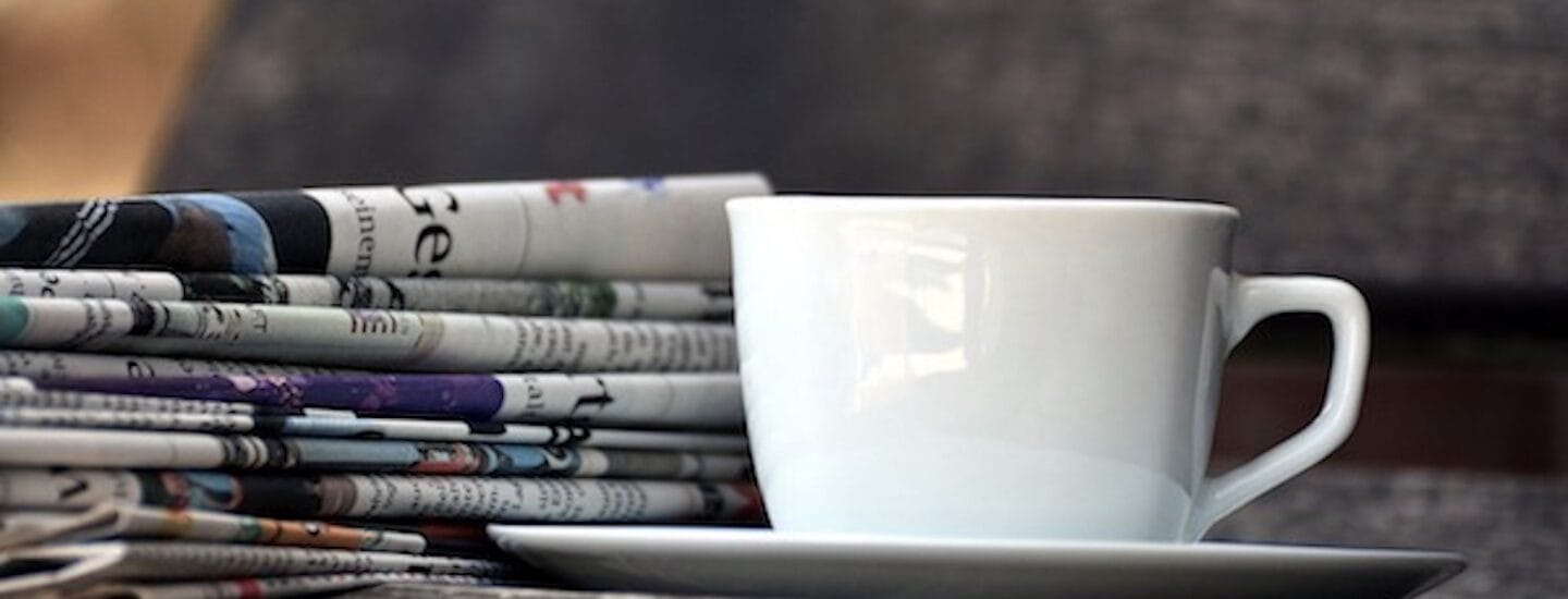 En hvid kaffekop står på en matchende underkop på en træbænk. Ved siden af den antyder en stak aviser, let foldet og slidt, at de er blevet læst. Denne rolige scene, fri for enhver tilknytningskrav, formidler ren afslapning og læseglæde.