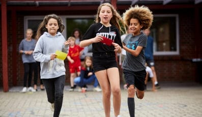 Tre børn løb glade udendørs og knugede farverige papirer på familie-sommerskolen. Den krølhårede dreng til højre har en blå skjorte, pigen i midten bærer sorte shorts og skjorte, mens barnet til venstre ifører sig en grå hættetrøje. Andre børn leger lystigt i baggrunden.