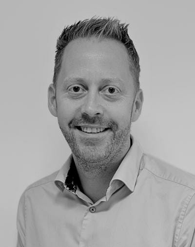 En mand med kort hår og ansigtshår smiler til kameraet. Han er iført en lys skjorte med knapper. Billedet er i sort/hvid.