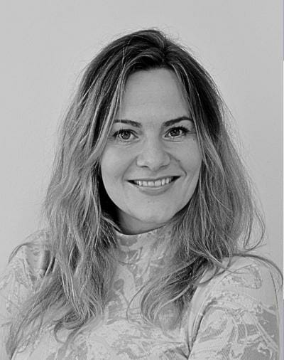Smilende person med langt hår iført en mønstret top, ser på kameraet i et sort-hvidt portræt.