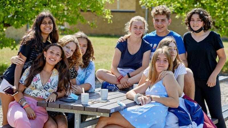A group of nine young people sits and stands casually around a picnic table outdoors, smiles wide as they enjoy the sunny day. With trees and a building in the background, its a scene reminiscent of those shared on the Danes Worldwide website, radiating a relaxed and cheerful atmosphere.