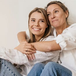 En ung kvinde og en ældre kvinde sidder tæt sammen og smiler varmt. Den unge kvinde hviler hovedet på den andens skulder. Begge er iført afslappede hvide skjorter og jeans, siddende mod en almindelig lys baggrund.