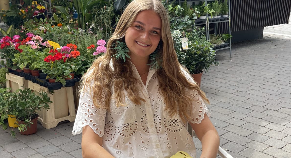 En ung kvinde med langt, bølget hår sidder på en stol uden for en blomsterhandler. Hun er iført en hvid blondekjole og har en gul vævet clutch. Bag hende står forskellige potteplanter og blomster. Belægningen er udført i grå mursten.