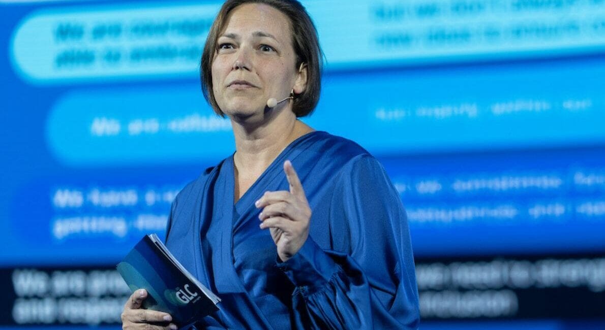 En kvinde i en blå bluse holder en notesbog og gestikulerer med højre hånd, mens hun taler på scenen. Baggrunden viser sløret tekst på en blå skærm, der foreslår en professionel eller konferenceindstilling.