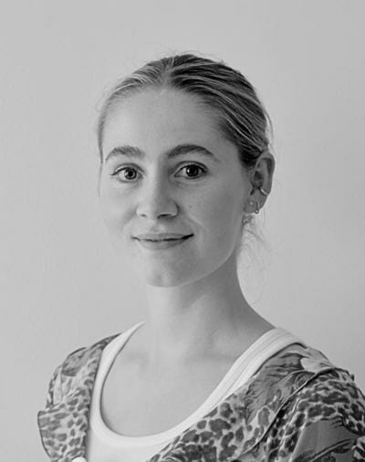 Et sort-hvidt portræt af en person med lyst hår trukket tilbage, iført en blomstermønstret top over en hvid skjorte. De vender let til siden med et neutralt udtryk mod en almindelig baggrund.