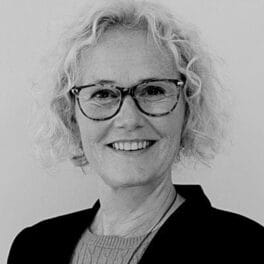 Sort/hvid foto af en smilende person med krøllet, kort hår og briller, iført en sweater og en blazer.