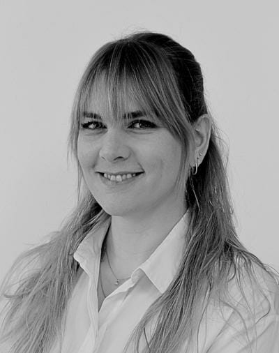 Sort og hvidt portræt af en person med langt, lyst hår og pandehår, smilende. De er iført en skjorte med krave og små øreringe. Baggrunden er almindelig.