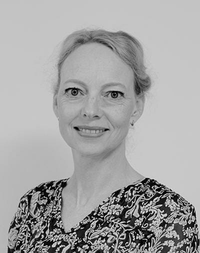 Et sort/hvidt portræt af en smilende person med lyst hår stylet op. De er iført en mønstret top med blomstermotiver og små øreringe, poseret mod en almindelig baggrund.