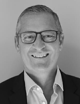 Et portræt i gråtoner af en smilende person iført briller, en jakkesæt og en skjorte med krave, med kort hår, mod en almindelig baggrund.