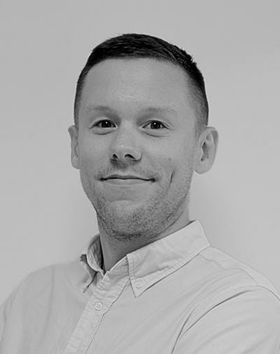 A person with short hair smiles slightly, wearing a light-colored collared shirt, against a plain background. The image is in black and white.