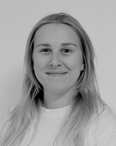 En person med langt hår smiler let til kameraet. De er iført en lys skjorte og små øreringe. Billedet er i sort/hvid.