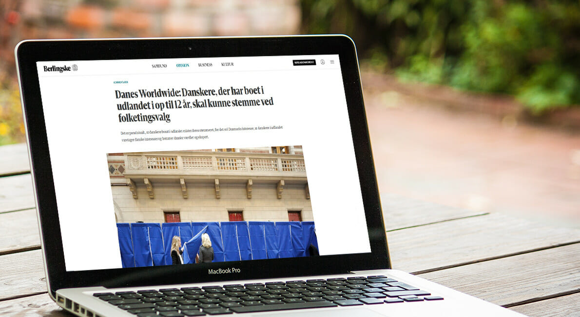 En bærbar, der viser en artikel på sin skærm. Overskriften nævner danskere, der har boet i udlandet i 12 år og stemt ved valg. Billedet på skærmen viser folk bag stemmebokse. Den bærbare computer står på et træbord udendørs.