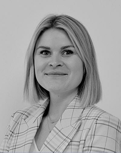 Sort og hvidt portræt af en person med skulderlangt hår, iført en plaid blazer og en halskæde. De kigger på kameraet med et lille smil på en almindelig baggrund.