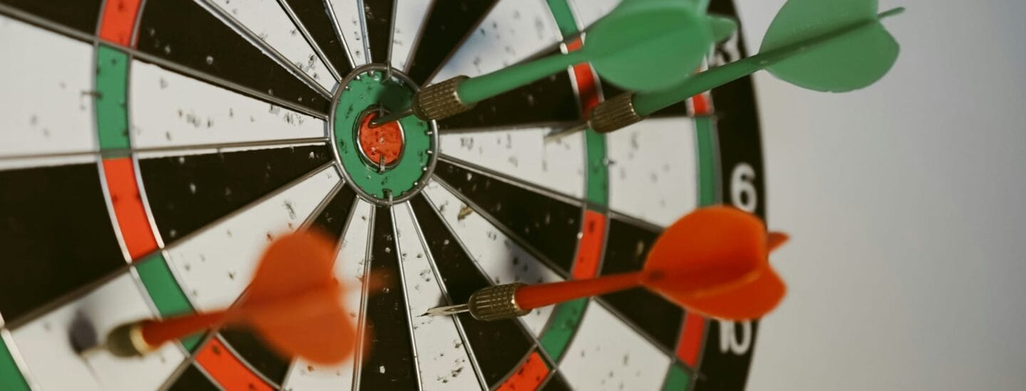 Nærbillede af en dartskive med fire pile. Tre grønne pile og en orange pil er indlejret i brættet. Den orange pil er tættest på bullseye, som er omgivet af et grønt og rødt cirkulært mønster.