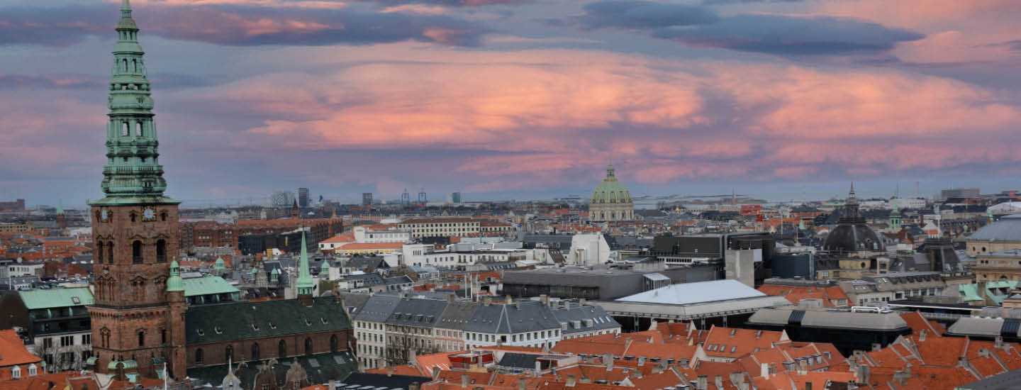 En panoramaudsigt over et bybillede i skumringen udfolder sig med et gammelt kirketårn, røde hustage og en dynamisk lyserød og lilla himmel. Forskellige bygninger spreder sig i landskabet, mens horisontens silhuetter mod den farverige solnedgang – en invitation til at udforske med rabat på hotelovernatninger.