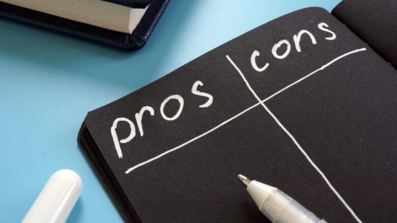 En notesbog med en sort side med en håndtegnet liste over fordele og ulemper. Ordene "pros" og "cons" er skrevet øverst, divideret med en lodret linje. En hvid pen og en delvist synlig bog ligger på en blå overflade ved siden af notesbogen.