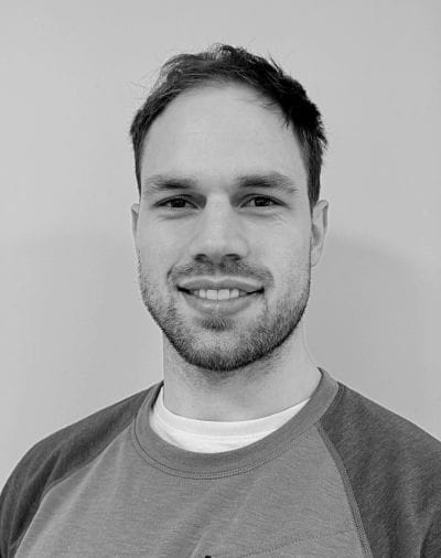 Black and white portrait of a person with short hair and a beard, smiling slightly. They are wearing a two-tone sweatshirt over a plain T-shirt, positioned in front of a plain background.