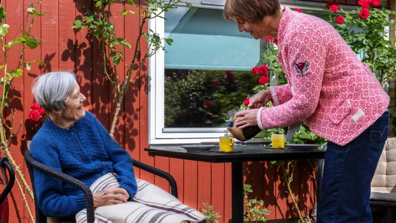 En kvinde hælder te i et krus til en ældre kvinde, der sidder i en stol uden for et rødt hus. De er omgivet af blomstrende blomster og grønt. Den ældre kvinde er dækket af et tæppe og iført en blå sweater, smilende mod den anden kvinde.