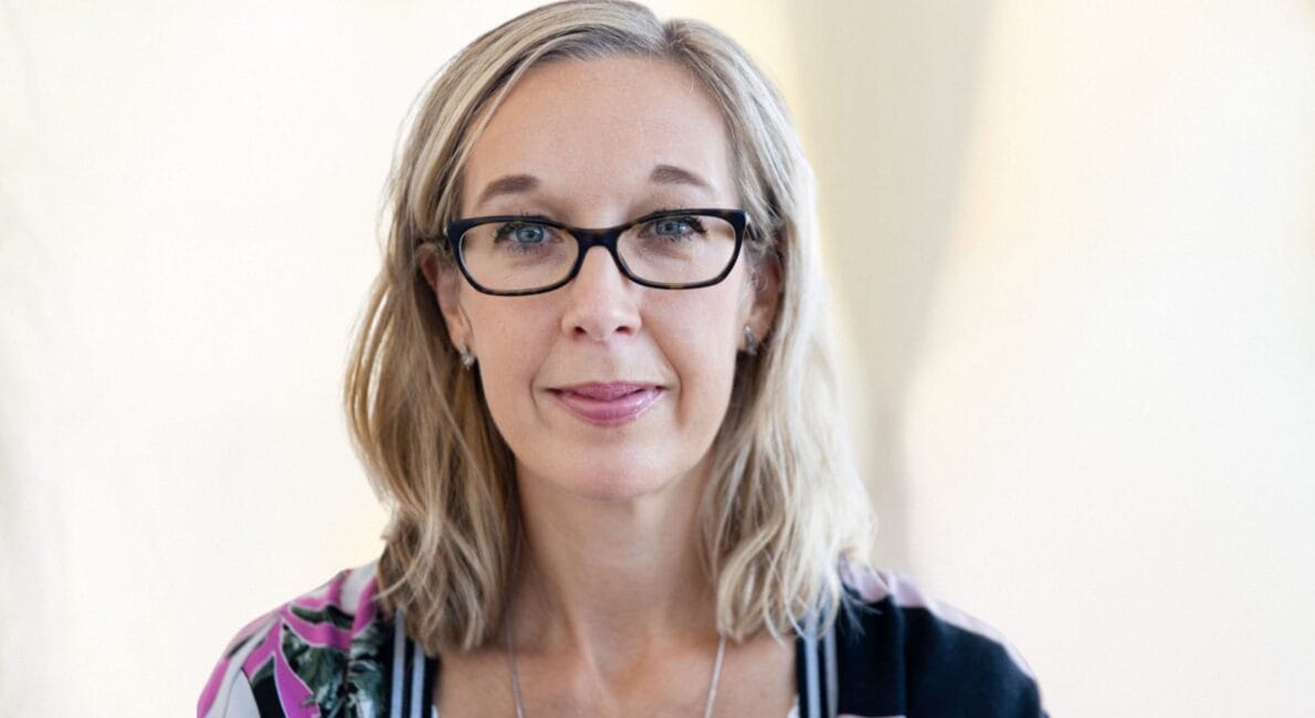 En person med skulderlangt blondt hår og briller ser på kameraet med et neutralt udtryk. De har en mønstret top og en halskæde på. Baggrunden er blødt sløret.