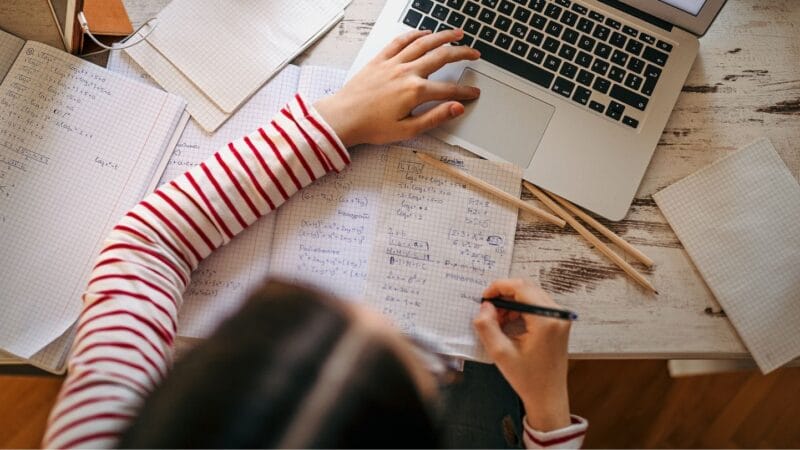 En person iført en rød- og hvidstribet skjorte skriver noter i en notesbog fyldt med matematiske ligninger, mens han bruger en bærbar computer. Forskellige notesblokke og blyanter er spredt på træbordet.