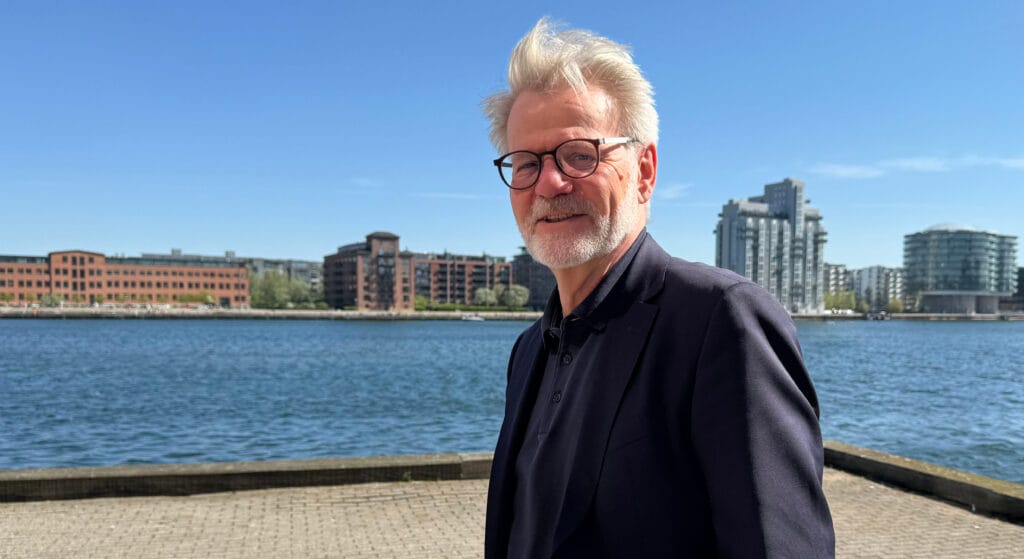 En ældre mand med hvidt hår og briller står ved en havnefront iført mørkt jakkesæt. Bag ham er moderne bygninger under en klar blå himmel.