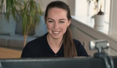En kvinde med langt brunt hår, iført en sort skjorte, smiler, mens hun deltager i et Danes Worldwide webinar på sin computer. Et webcam er placeret på skærmen. Bag hende er det en hyggelig sofa, levende plante og store vinduer, der sætter scenen.