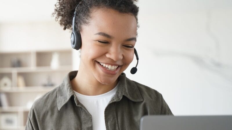 En kvinde med krøllet hår smiler, mens hun bærer et headset. Hun ser på en bærbar computerskærm i et lyst rum med hylder i baggrunden. Hun fremstår engageret og tilfreds.