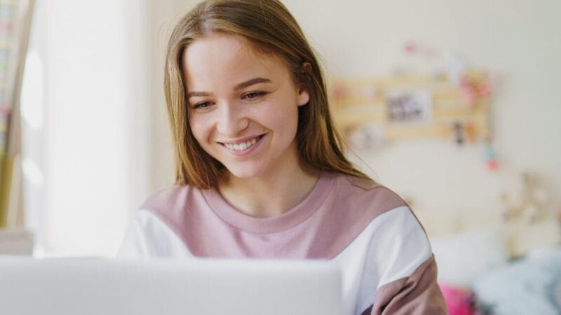 En ung kvinde med langt hår og et smil ser på en bærbar skærm. Hun er iført en pink og hvid sweatshirt i et hyggeligt værelse med slørede pynteting i baggrunden.
