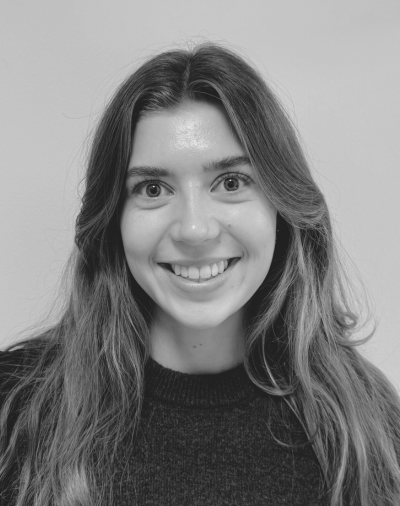 A person with long hair smiling at the camera, wearing a dark sweater against a plain background. The image is in black and white.