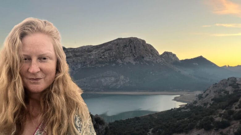 En person med langt blondt hår står foran et naturskønt bjerglandskab ved solnedgang. Bag dem er der et roligt vandområde og forrevne bakker under en farverig himmel, der skifter fra dag til nat.