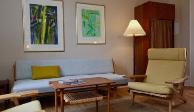 En hyggelig stue med en grå sofa, gul pude og et sofabord i træ. To abstrakte malerier hænger over sofaen. En gul lænestol og en høj gulvlampe tilføjer varme til rummet. Et skab med et gardin er i hjørnet.