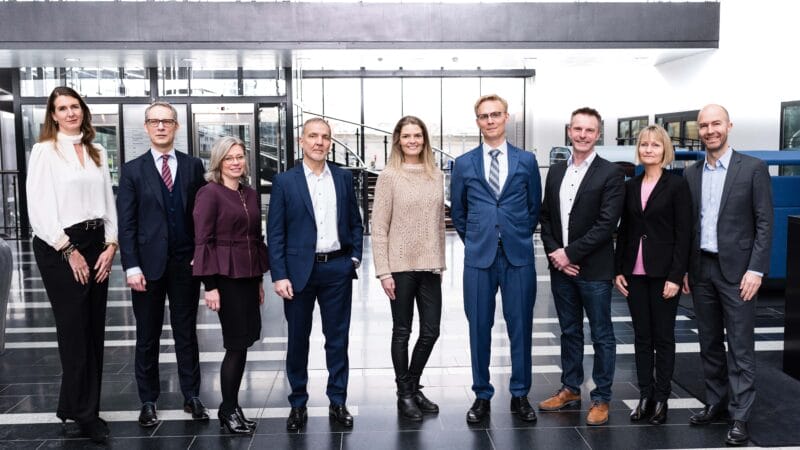Ni eksperter står i en kontorlobby, alle i professionel påklædning. Arrangeret i en linje og vendt mod kameraet, er de sat på baggrund af vinduer og en trappe. Scenen fuldendes af det slående sort-hvide klinkegulv.