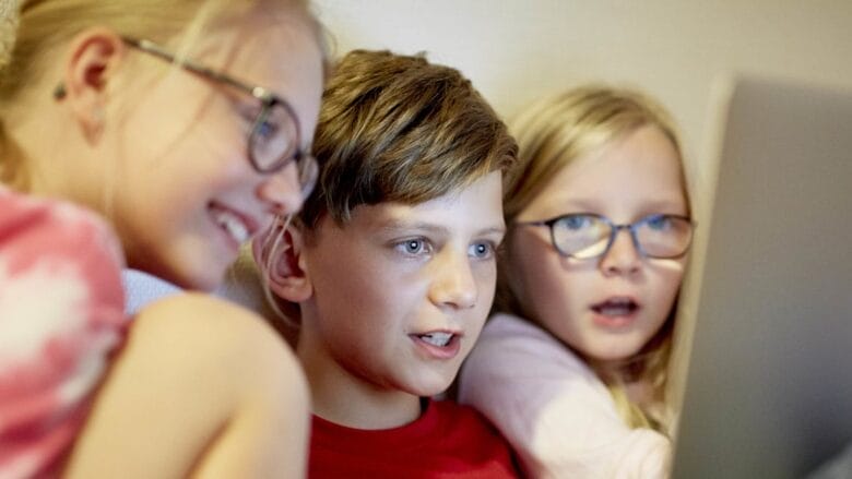 Three children are sitting closely together, intently watching an online video on a laptop screen. They appear engaged and interested, with two of them wearing glasses. The scene suggests they are either being entertained or learning something new in the digital world.