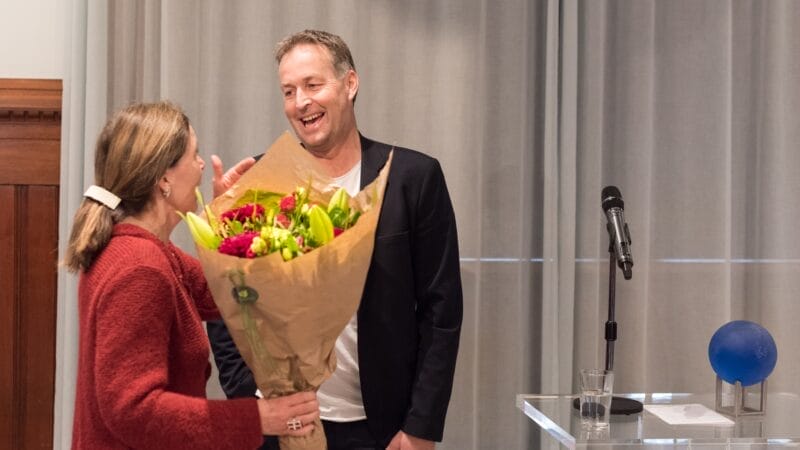 En kvinde i en rød frakke rækker en buket blomster til en smilende mand i sort jakkesæt. De står ved siden af et klart podium med en mikrofon og en blå kugle på. Indstillingen ser ud til at være indendørs, muligvis ved et arrangement.