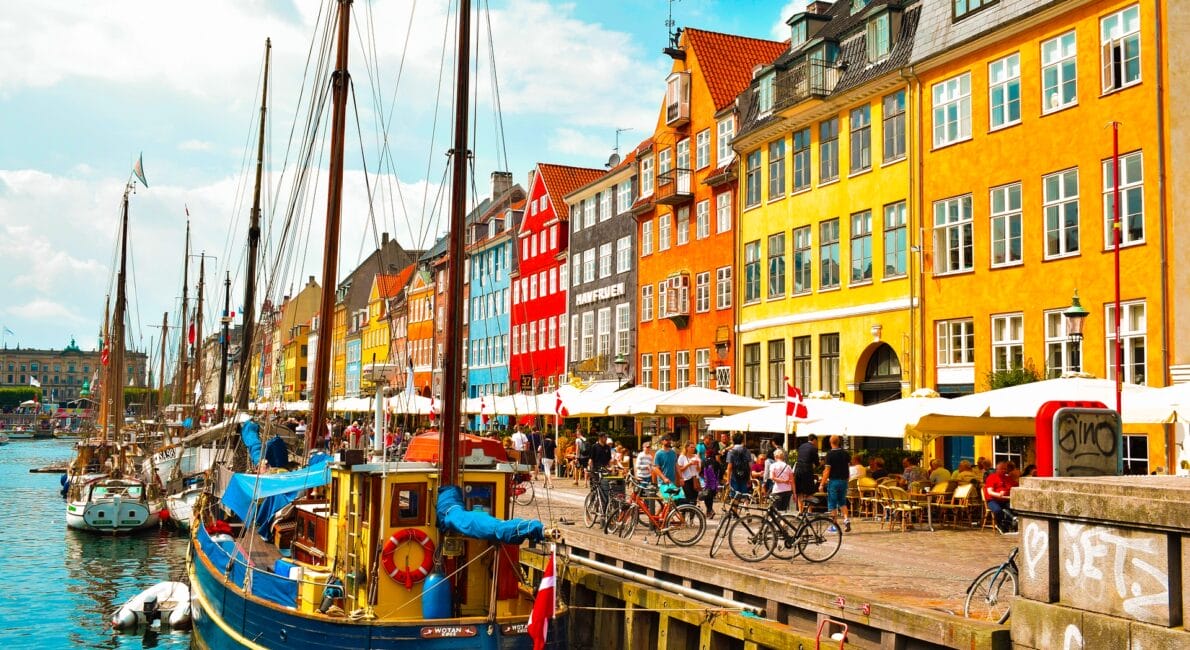 En livlig scene ved vandet med farverige historiske bygninger i rød, gul og orange. Fortøjede både ligger langs vandet, og folk nyder udendørs spisning på caféer langs brostenspromenaden under en delvist overskyet himmel, der minder om den charme, der fejres af dem med Dansk statsborgerskab ved fødslen.