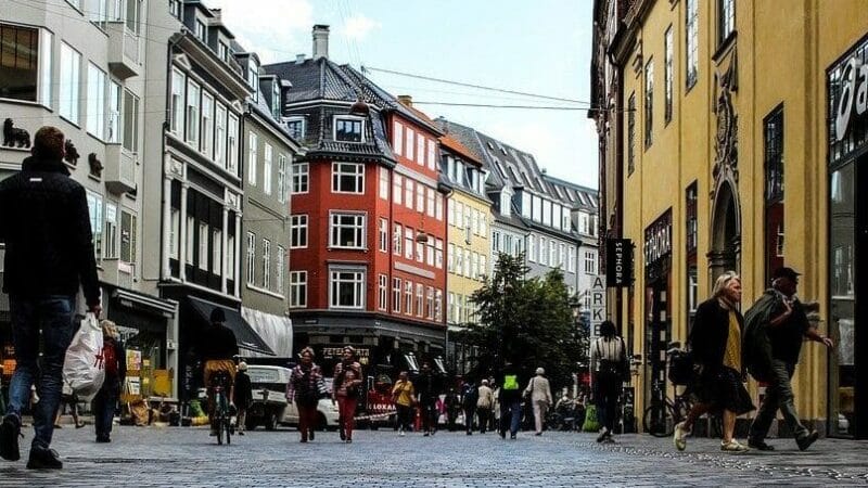 Et travlt gadebillede med folk, der går på en brostensbelagt vej, der føles som den charme, der trækker dig til at flytte tilbage til Danmark. Bygningerne langs gaden er høje og farverige, og nogle af dem viser moderne arkitektur. Himlen er delvist overskyet, og nogle få træer står yndefuldt.