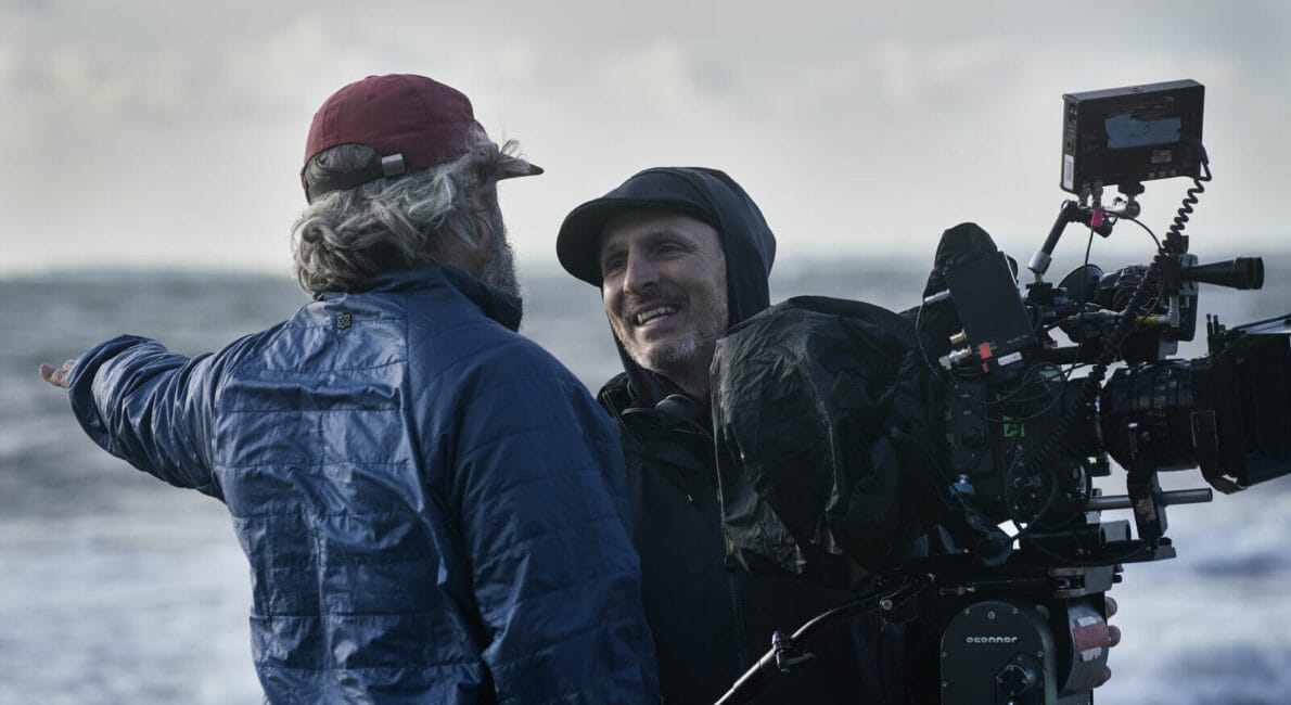 To mennesker ved havet er engageret i samtale. Den ene, iført rød kasket og blå jakke, gestikulerer udad. Den anden, i sort hat og jakke, betjener et kamera på et stativ. Havet og overskyet himmel danner baggrunden.