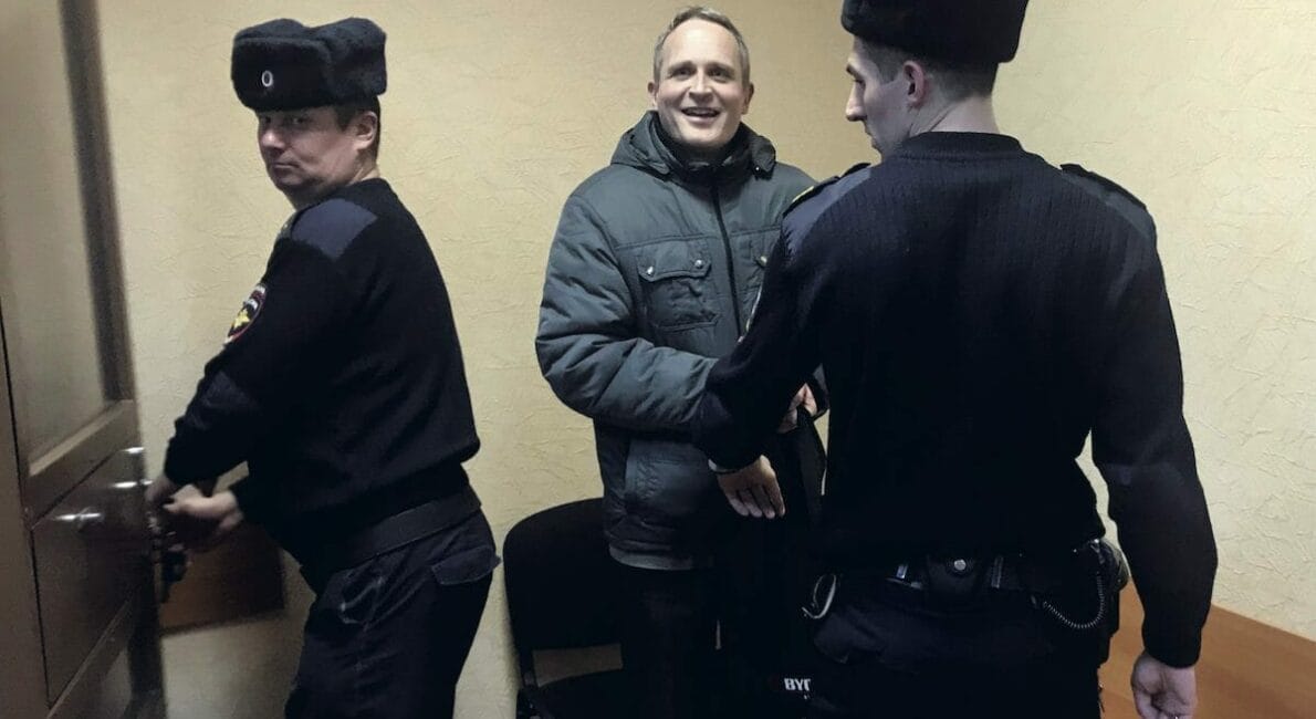En mand i en grå frakke smiler, mens han står mellem to uniformerede betjente i et værelse. Den ene betjent åbner en dør, og den anden holder i mandens arm. Rummet har almindelige vægge og træbænke.