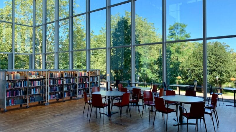 Et rummeligt bibliotek med store glasvinduer med udsigt over en frodig, grøn udendørs scene. Indenfor er der bogreoler fyldt med bøger og flere runde borde omgivet af røde stole på et trægulv. Naturligt lys fylder rummet.