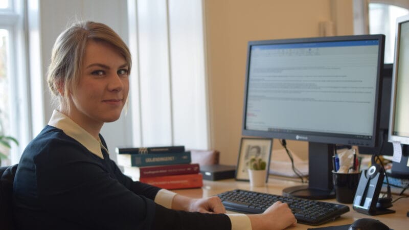 En kvinde i en mørk blazer sidder ved et træskrivebord og arbejder på en computer. Hun kigger mod kameraet med et lille smil. Der er bøger stablet på skrivebordet og et indrammet foto ved siden af skærmen. Naturligt lys fylder rummet.