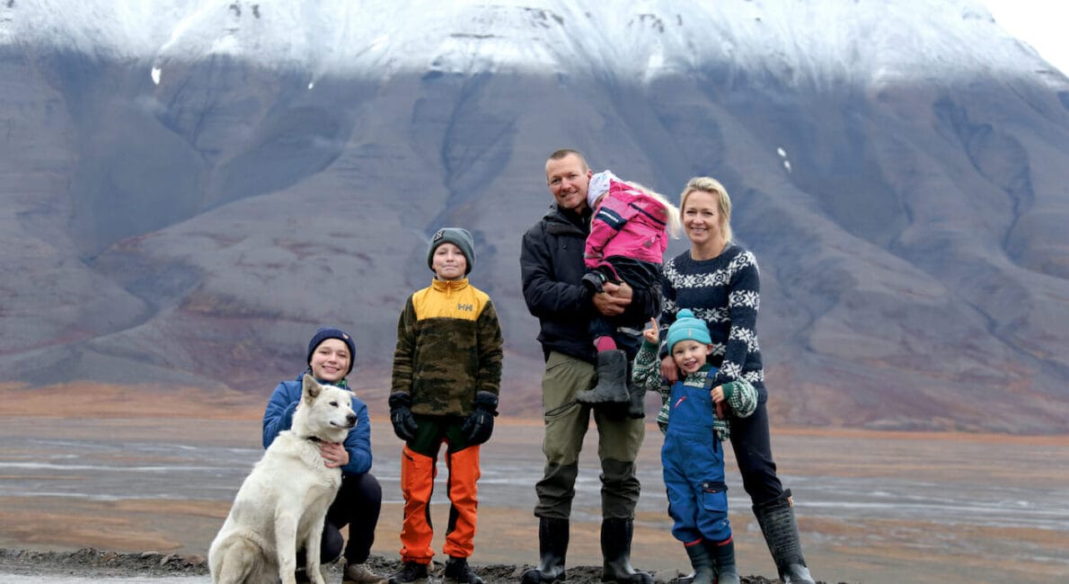 En familie på fem står på en vej med en hvid hund, foran snedækkede bjerge. Faderen holder et barn, mens et andet barn står ved siden af moderen. De er alle klædt varmt på, hvilket tyder på et koldt klima.