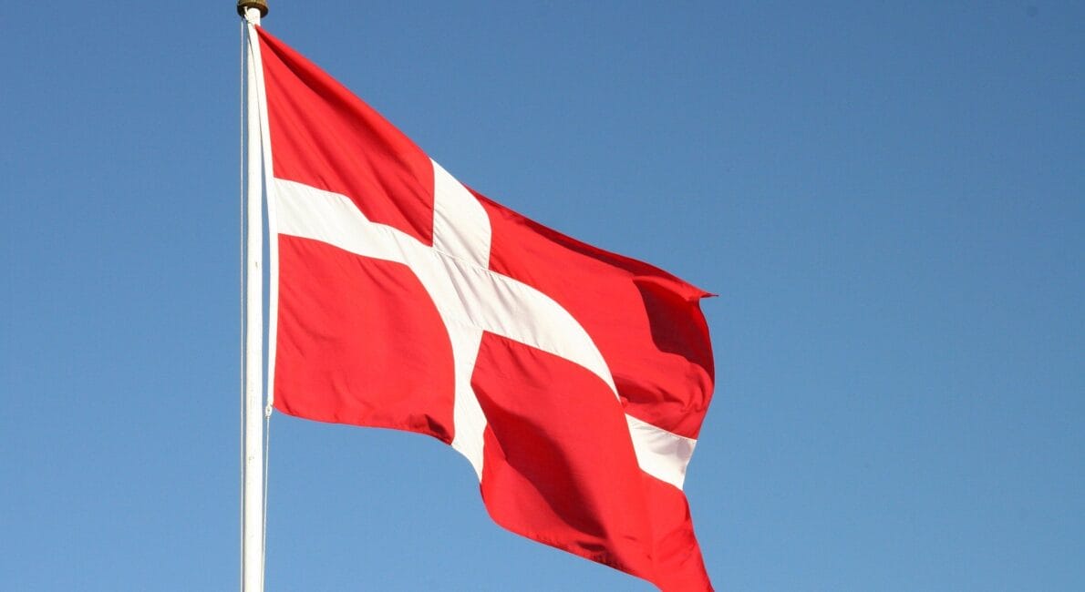 Billedet viser Danmarks flag, der stolt vajer på en flagstang under en klar blå himmel, som symboliserer national identitet under diskussioner om "betingelser for generhvevelse af dansk statsborgerskab." Det røde flag med sit hvide skandinaviske kors står levende på den azurblå kulisse.