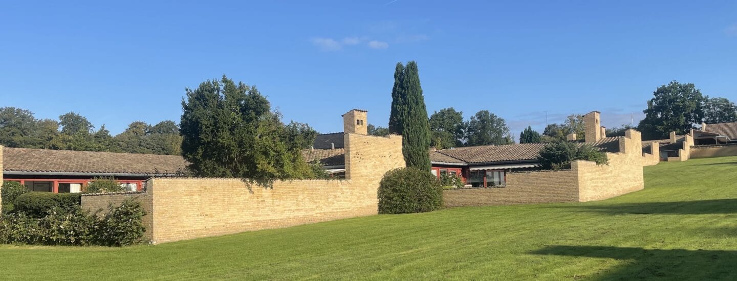 A scenic view of a brick building complex with sloped roofs, surrounded by lush green grass and trees. The architecture features several chimneys and the sky is clear and blue.