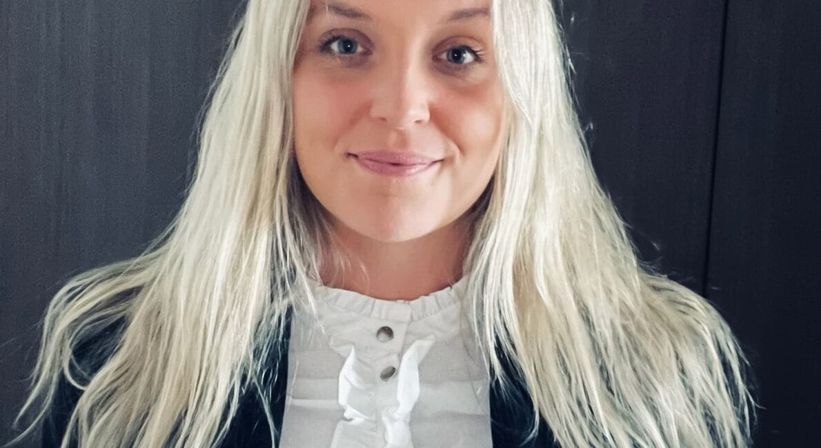 En person med langt blondt hår smiler let. De har en sort blazer på over en hvid flæsebluse. Baggrunden er en almindelig mørk overflade.
