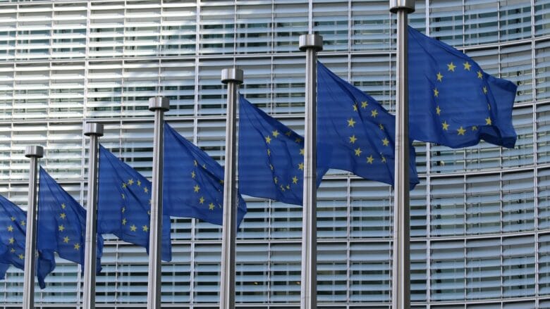Seks EU-flag med gule stjerner på blå baggrund står opstillet, blafrende i vinden. De er sat op mod en moderne, gittermønstret bygningsfacade.