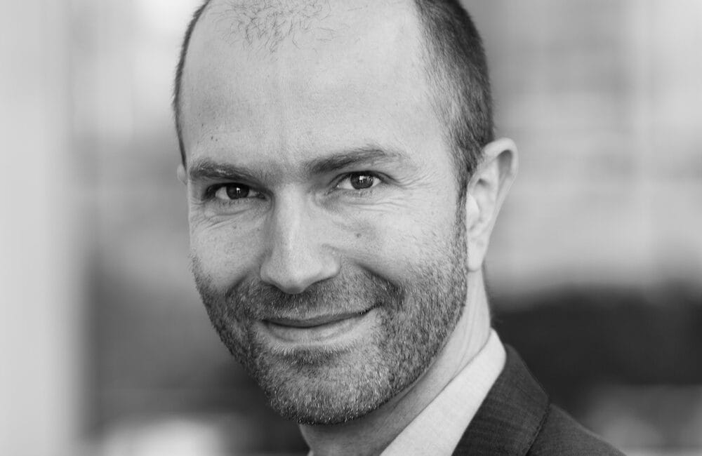 En mand med skæg og et venligt smil er iført jakkesæt. Baggrunden er sløret, hvilket tyder på en indendørs indstilling med store vinduer. Billedet er i sort/hvid.