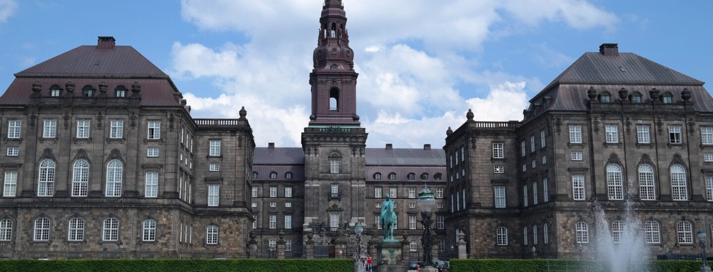 Historisk bygning med et centralt tårn og symmetriske fløje med klassisk arkitektur. En statue er foran omgivet af en bred plads og et springvand. Blå himmel og skyer i baggrunden.
