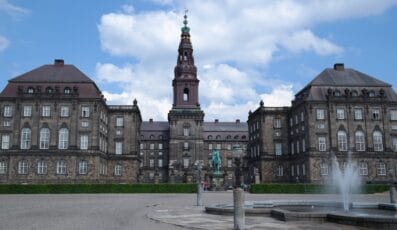 Historisk bygning med et centralt tårn og symmetriske fløje med klassisk arkitektur. En statue er foran omgivet af en bred plads og et springvand. Blå himmel og skyer i baggrunden.